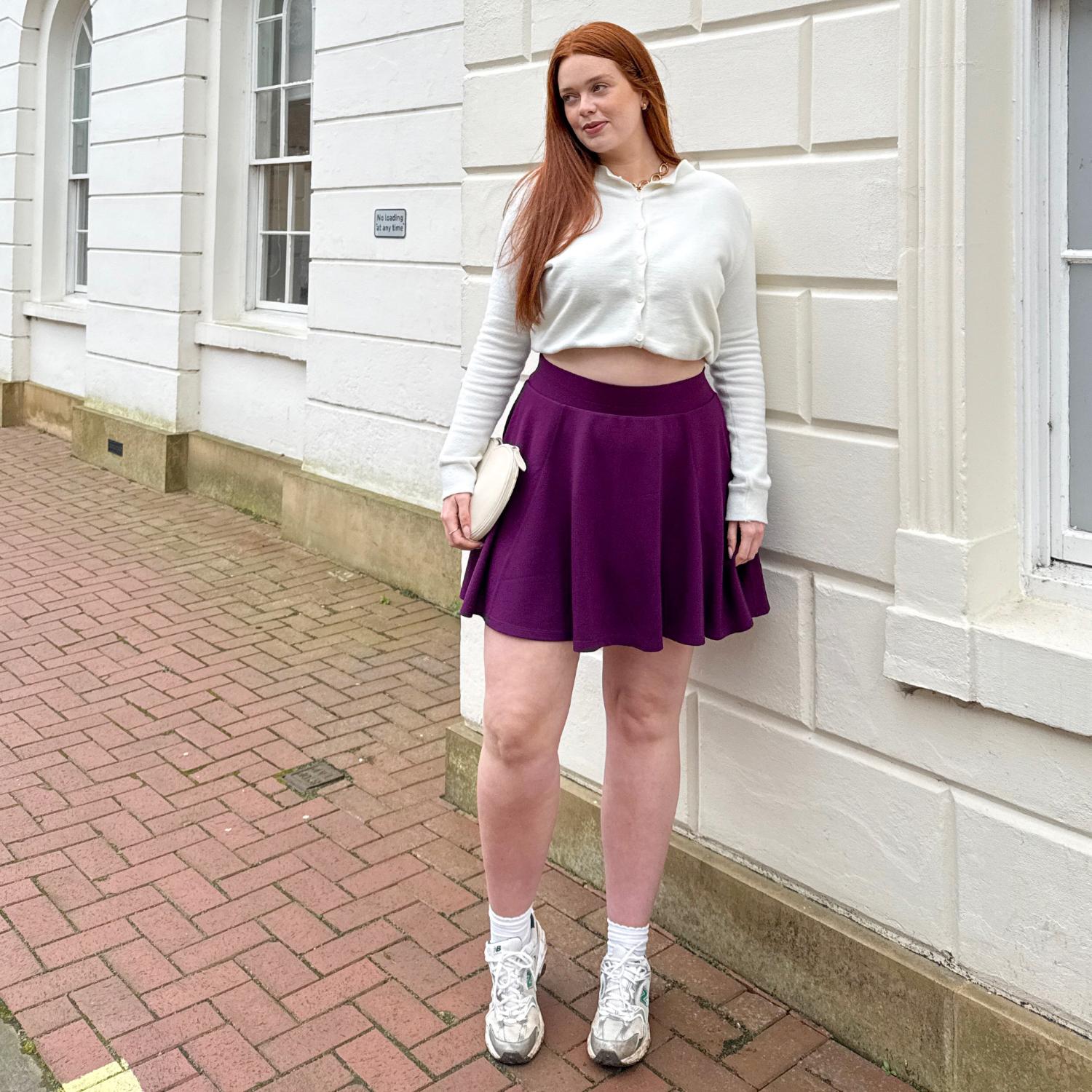 Woman in a white crop top and purple skirt standing on a brick sidewalk next to a white building.
